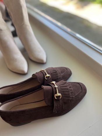 Loafer Geanne bruin met goudkleurige gesp, naast witte enkellaarzen op vensterbank