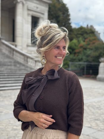 Vrouw draagt bruine sweater met strikdetail over Lizalau satin dress taupe, buiten op terras met klassieke architectuur op ac