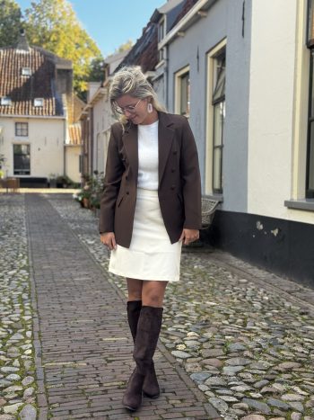 Vrouw in zwarte blazer, wit topje en witte rok met zwarte laarzen loopt over kinderkopje straat.