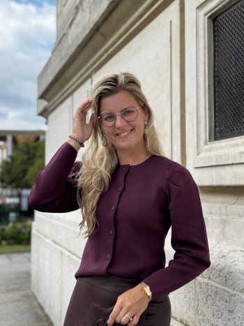 Vrouw met blonde haren en bril draagt paarse blouse buiten voor beige gebouw