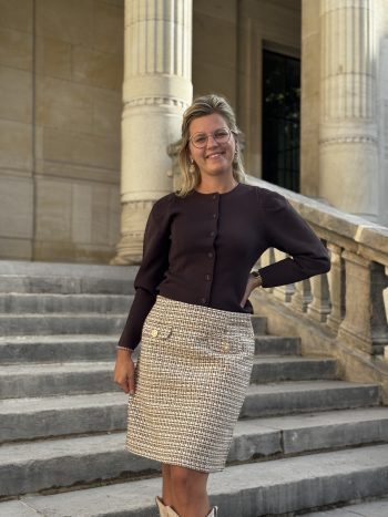 Vrouw in zwarte cardigan en taupe rok met fijnmazig patroon op stenen trappen voor klassiek pand met zuilen